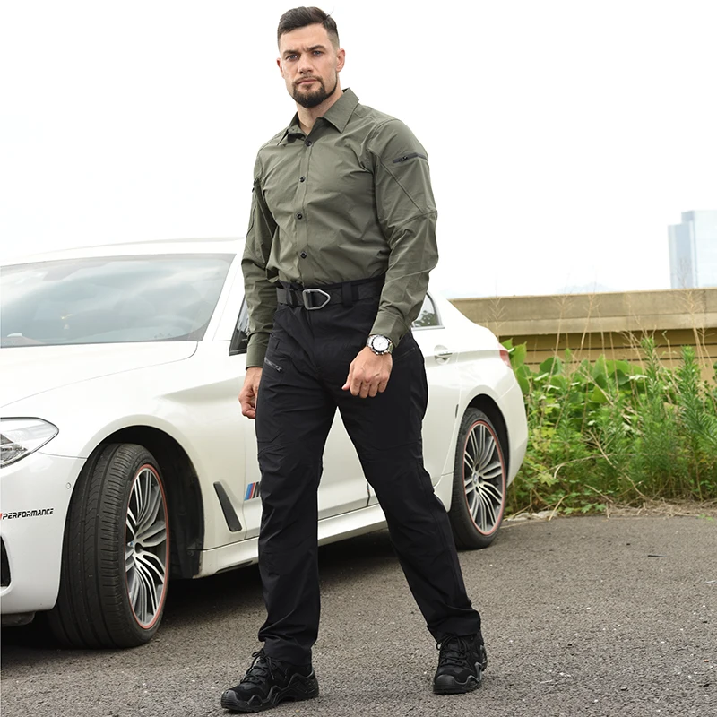 Un hombre que usa una camisa verde y pantalones negros se encuentra frente a un coche blanco.