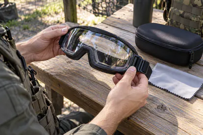 Manos revisando unas gafas cerradas de protección sobre una mesa junto a su funda